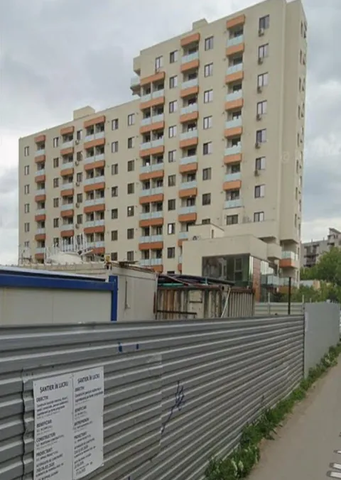 Fotografie a blocului de locuințe Emil Palade din Iași, finalizat, văzut de pe stradă, cu un gard de șantier în prim-plan